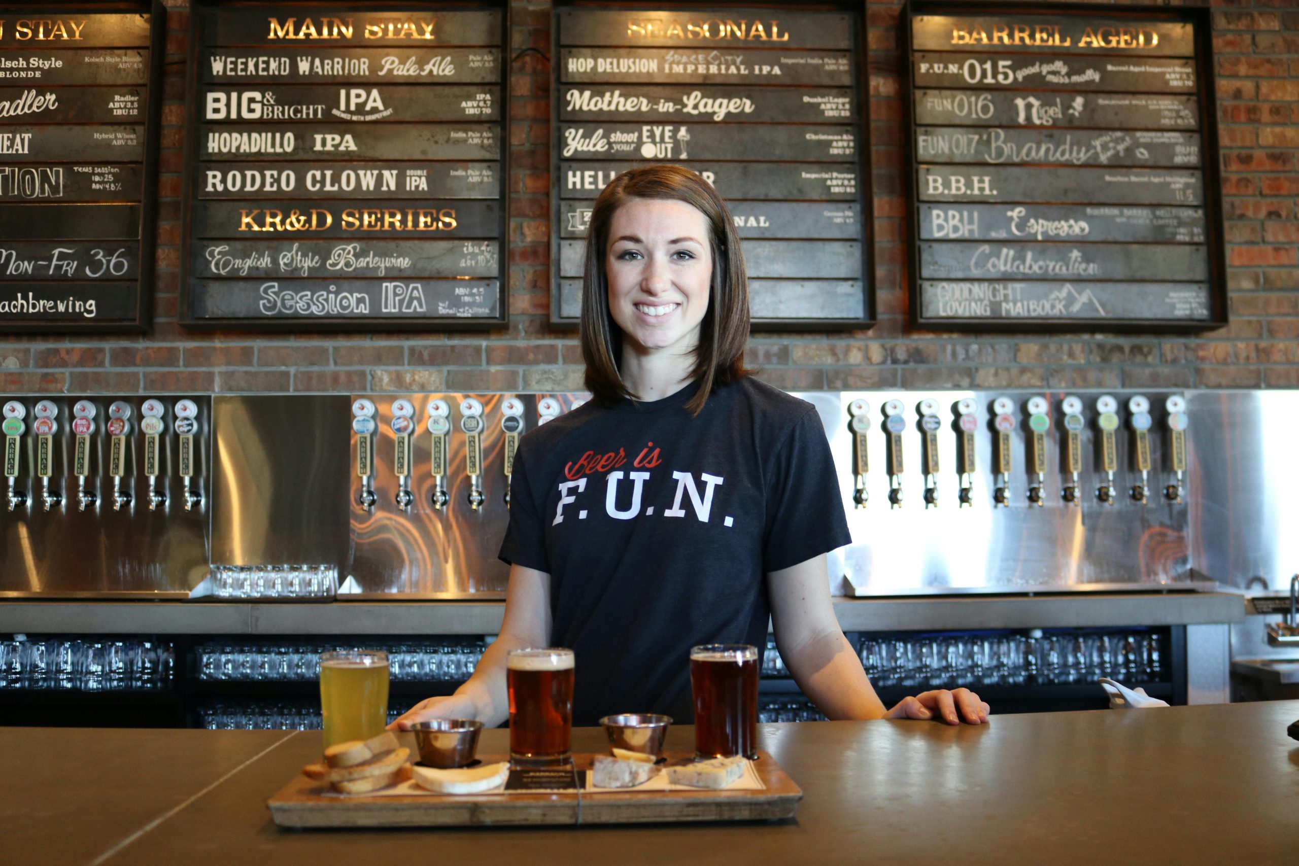 Services Smiling bartender in a bar serving craft beer flights with taps and menu board in the background.