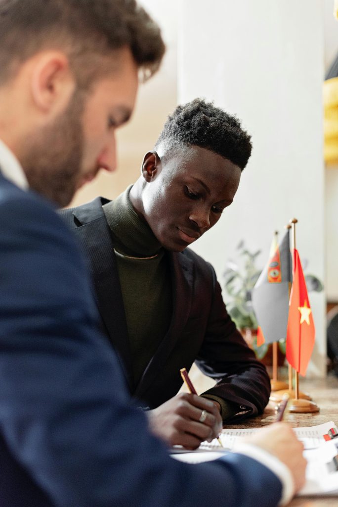 Two businessmen in formal wear signing an international agreement with flags on table.